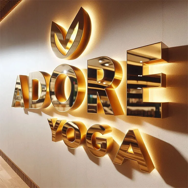 A picture of a Custom Backlit Sign mounted on a wall. The product says ADORE YOGA. The product is gold and illuminated.