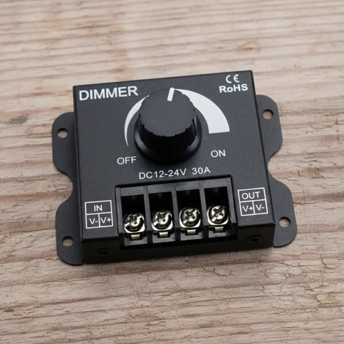 A black LED dimmer device is centrally positioned in a rustic, wooden workshop or "signs atelier" environment. The dimmer is labeled DC 12-24V 30A and features a large rotary knob (potentiometer) that turns between "OFF" and "ON." Screw terminal connectors for "IN" and "OUT" are visible on the right side of the device. The dimmer rests on a dark brown, distressed wooden workbench. Various tools and equipment related to handicrafts and sign-making are scattered yet organized on the bench and in the background: leather rolls, scissors, metal stencil letters, a brass plate engraved with "SIGNATELIER", brushes, jars of paint, and vintage-looking wooden signs and technical drawings hanging on the wall. The scene is illuminated with a natural, warm light.