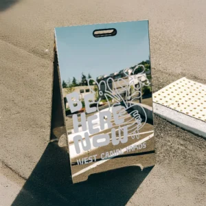 Mirrored chrome A-frame sidewalk sign featuring a floral illustration and white bold text reading 'BE HERE NOW' for West Canal Yards.