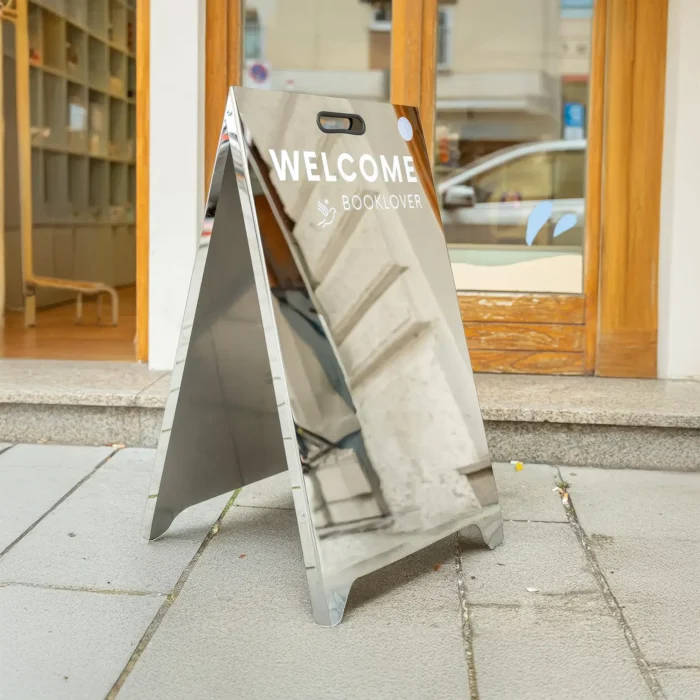 Mirrored sidewalk sign in a chrome A-frame design, featuring white 'WELCOME BOOKLOVER' text and a bird icon reflecting the street view.
